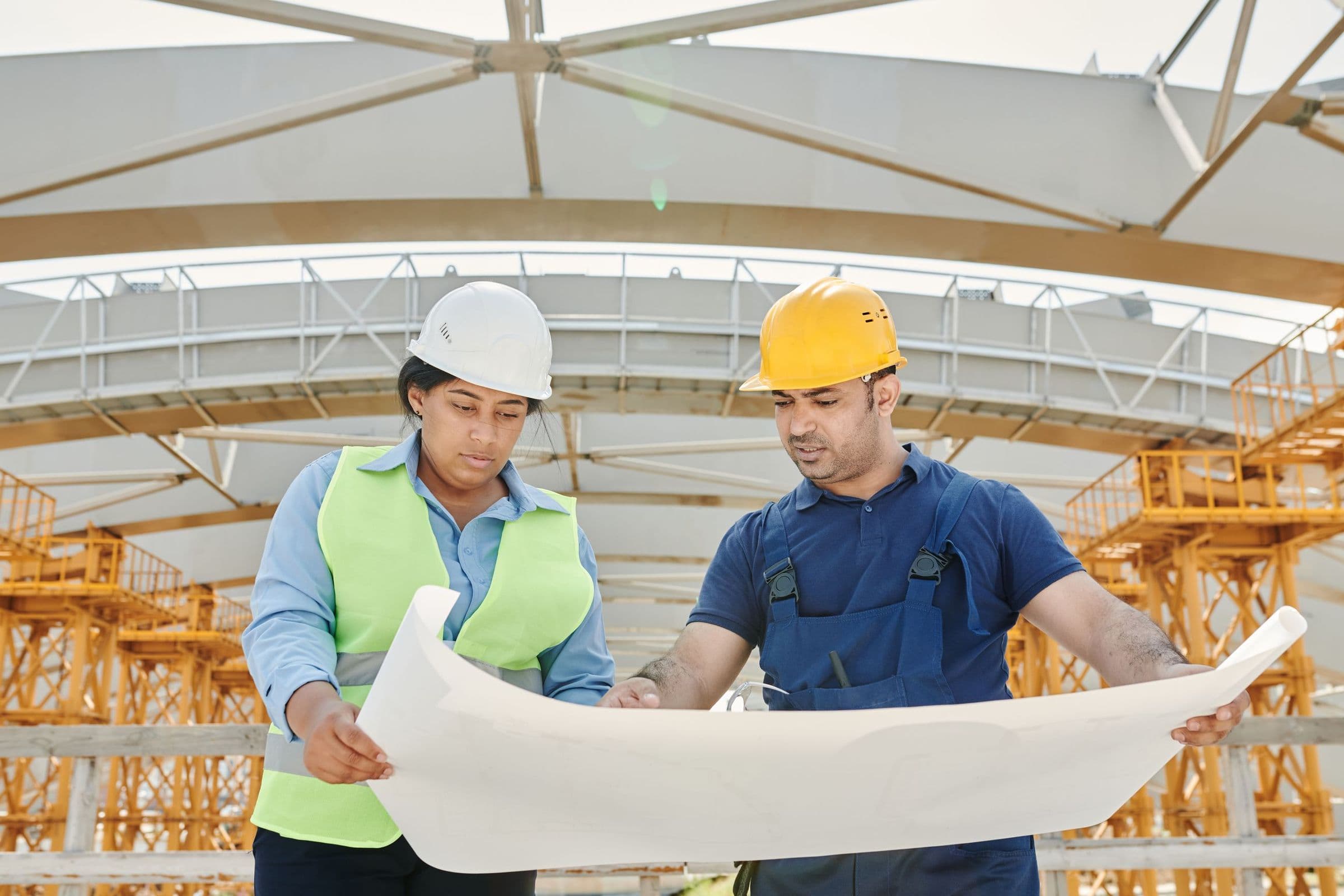 Two engineers in hard hats reviewing a large drawing beneath a steel roof structure