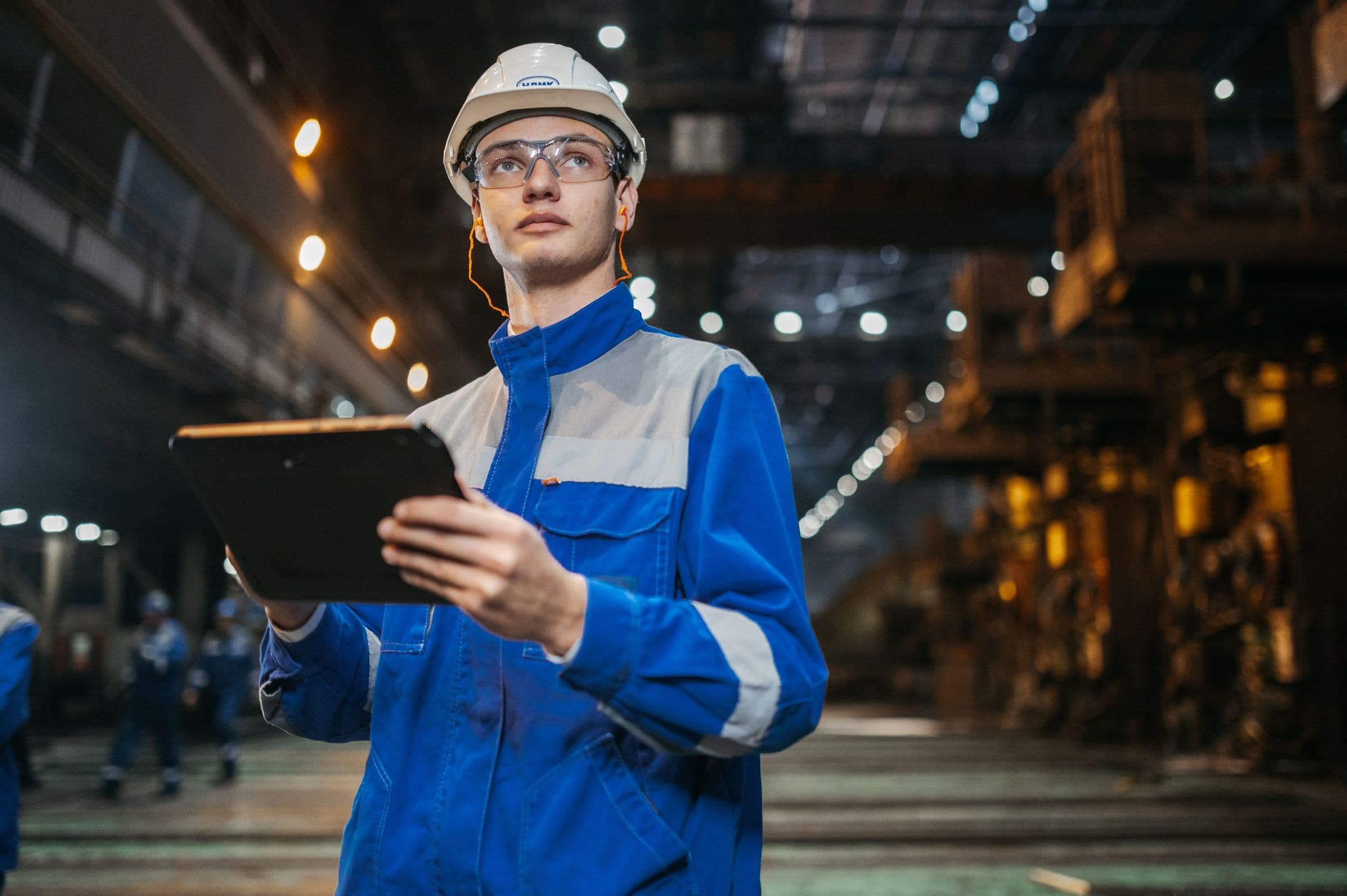 An engineer in a hard hat and safety glasses holding a tablet on a steelworks floor