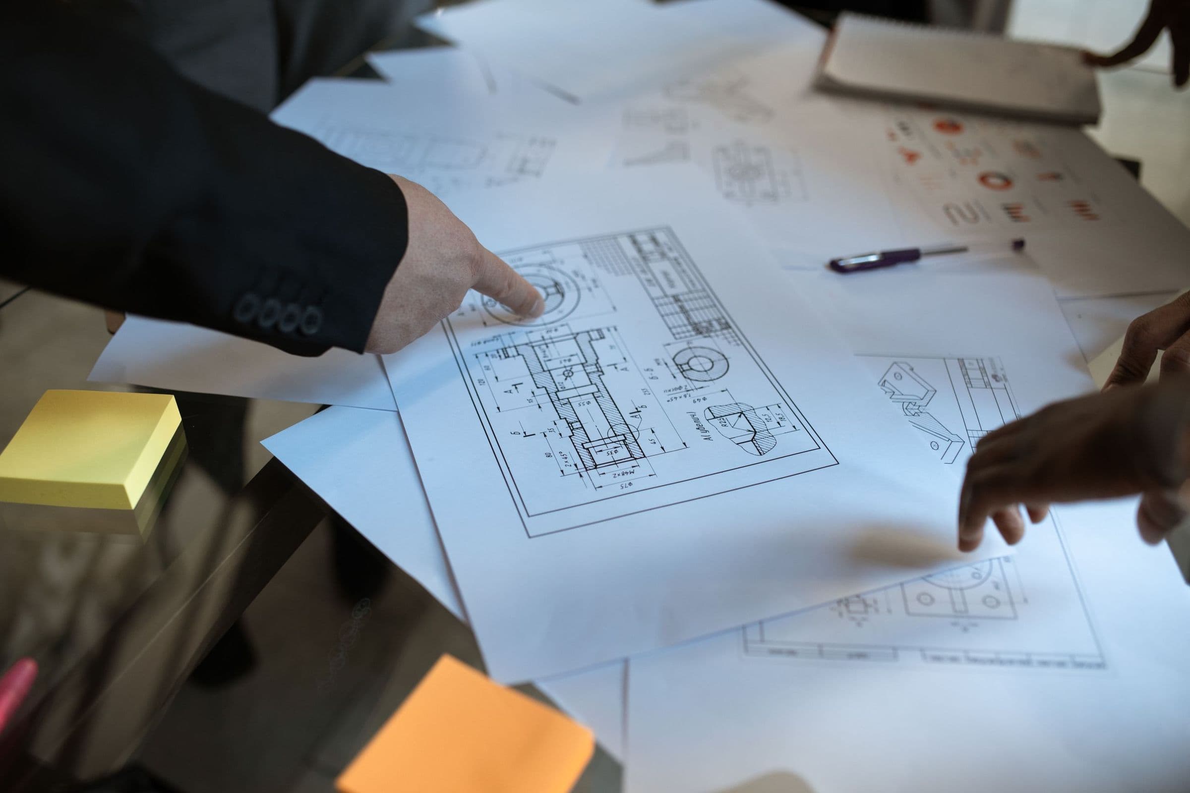Hands pointing out details on printed technical drawings spread across a desk